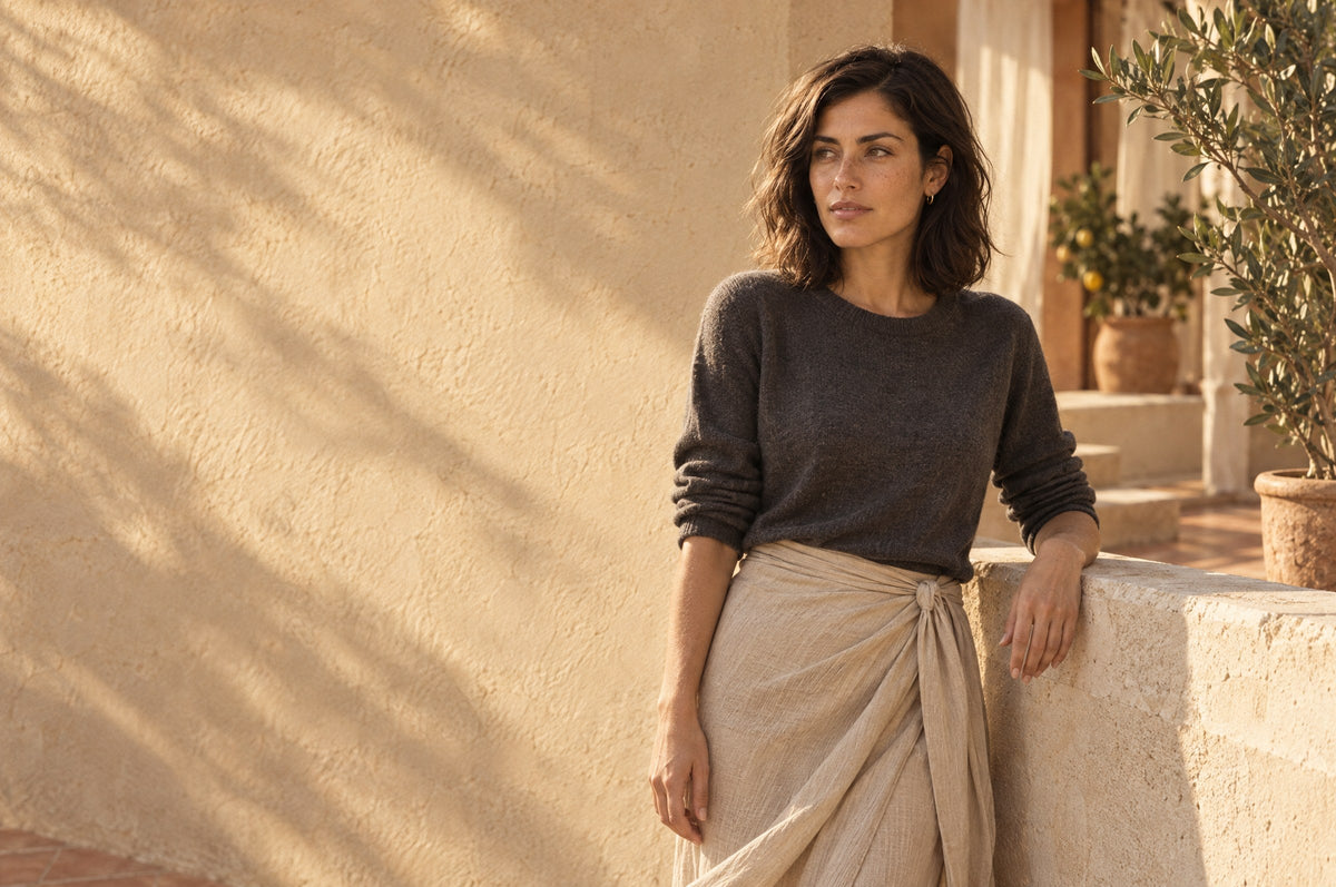 Woman in a dark knit and a long sand-colored wrap skirt leaning on a sunlit stone parapet