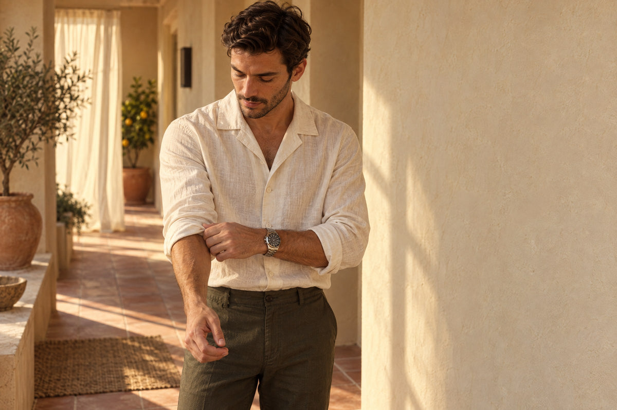 Man rolling up the sleeve of a white camp collar shirt in a sunlit hallway