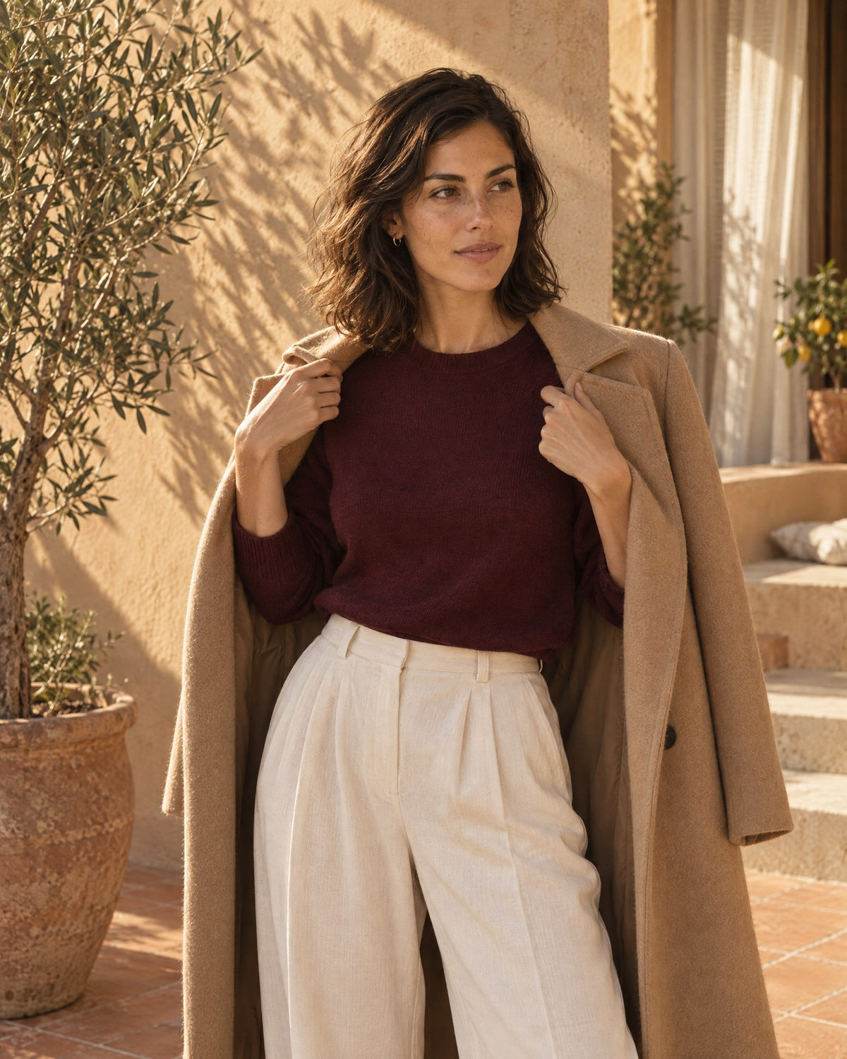 Woman slipping a long camel coat over her shoulders, worn with a deep bordeaux knit and wide cream trousers