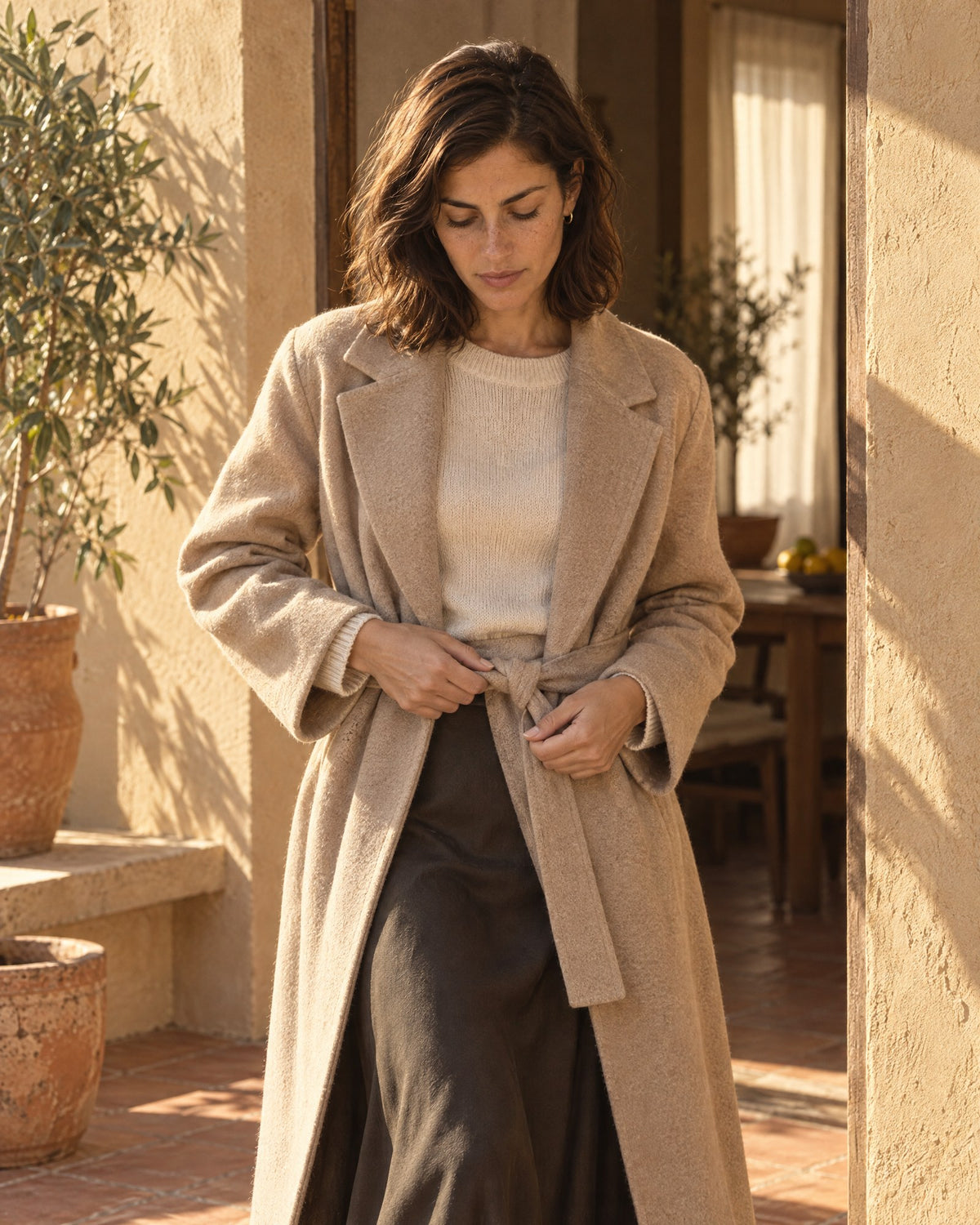 Woman fastening the belt of a long sand coat over a cream knit in a sunlit doorway