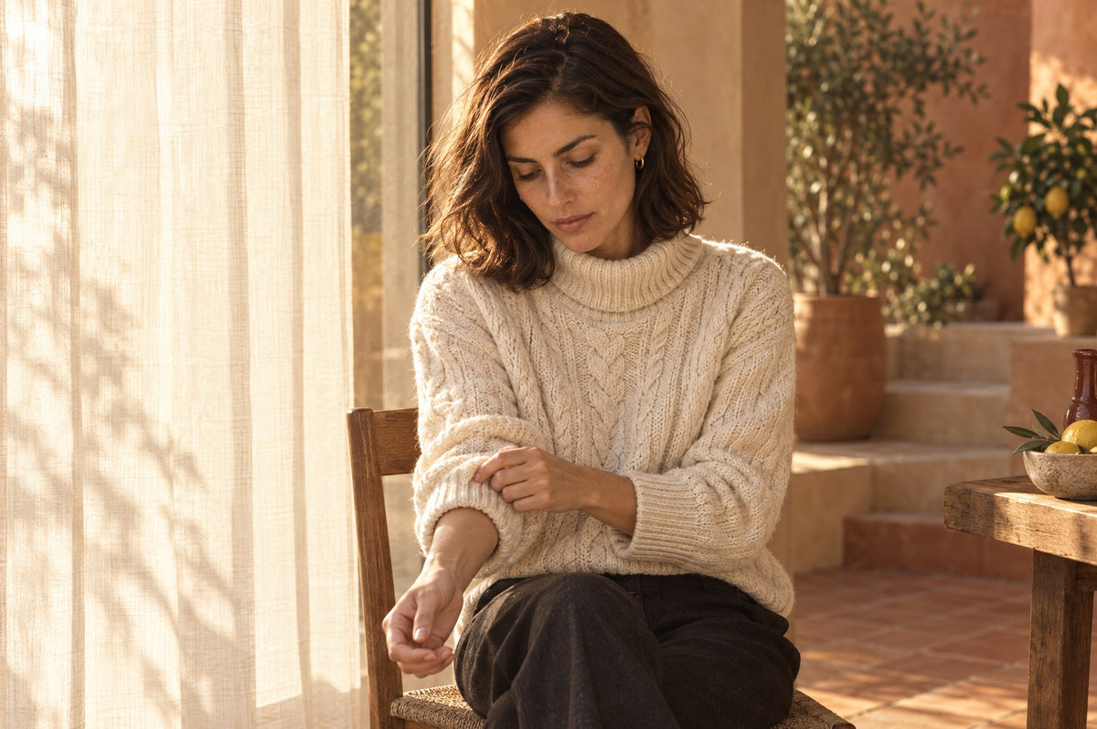 Woman in a cream cable knit sweater adjusting her cuff by a curtained window